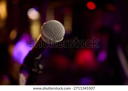 Stage microphone close-up ready for performance with shallow bokeh background, textured grille catching warm stage LEDs and blurred band equipment suggesting