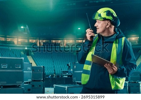 Stage supervisor in safety gear communicates via walkie-talkie while holding tablet during concert equipment setup in arena. Stage Equipment Installation.