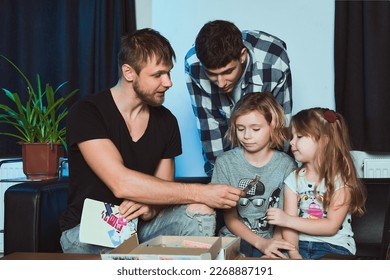 Staged photo  Homosexual couple   their children  two cute girls  at home   An everyday evening after day's work  Time to play and the kids  Let's see what's in the big box!                      