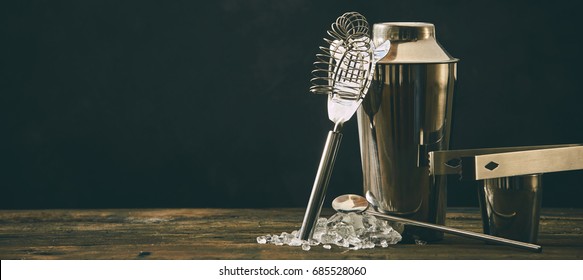 Stainless Steel Alcoholic Cocktail Bar Utensils, Ice And Equipment On A Rustic Wooden Bench Top Against A Black Background.