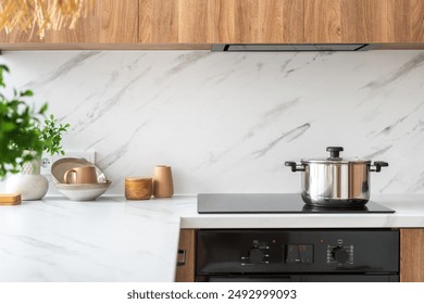 A stainless steel pot sits on a black electric stove top in a modern kitchen. Countertop are white marble and cabinets are light wood. Kitchenware and utensils on table with copy space - Powered by Shutterstock