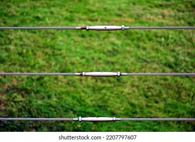 Stainless Steel Railing In Selective Focus. Modern Construction Materials. Shiny Steel Turnbuckle. Wire Cable Rope And Connection. Perspective View. Blurred Green Lawn Background. Ties And Connections