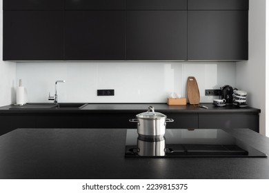 stainless steel saucepan with lid on induction stove on island countertop, contemporary cupboard, sink, water tap and kitchenware at stylish kitchen