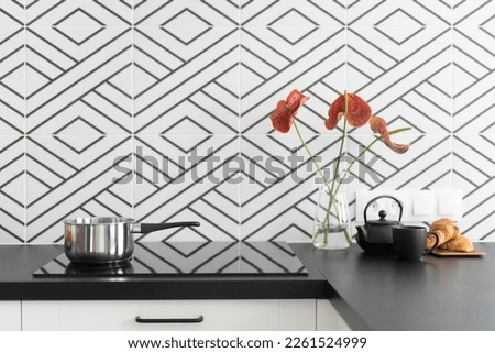 stainless steel saucepan on electrical stove, flowers in glass vase, fresh croissants on wooden plate, teapot and cup on countertop in kitchen with cozy interior