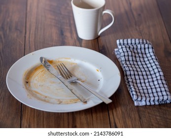 Stainless Steel Simple Knife And Fork On A Dirty White Plate With Napkin After Diner On A Wooden Table Surface. White Tea Cup And Classic Towel.