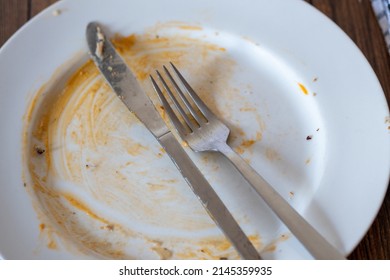 Stainless Steel Simple Knife And Fork On A Dirty White Plate With Napkin After Diner On A Wooden Table Surface.