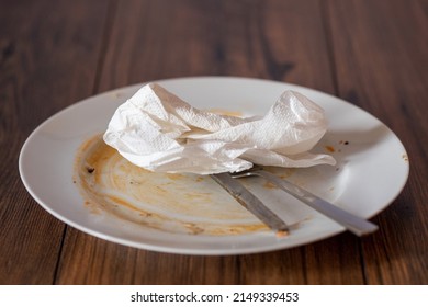 Stainless Steel Simple Knife And Fork On A Dirty White Plate With Napkin After Diner On A Wooden Table Surface.