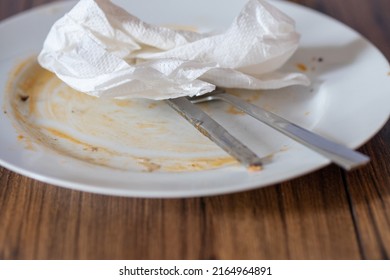 Stainless Steel Simple Knife And Fork On A Dirty White Plate With Napkin After Diner On A Wooden Table Surface.