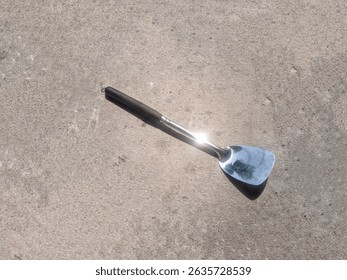 A stainless steel spatula or turner with a black handle, likely used for cooking or serving, resting on a textured concrete surface with a bright reflection on its blade. - Powered by Shutterstock - Get 15% off with code: PIKWIZARD15