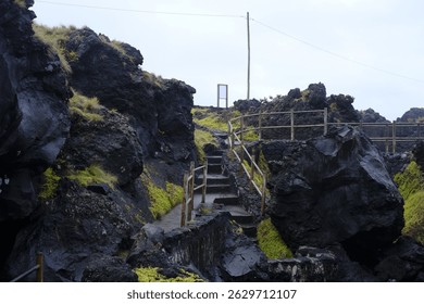 A stairway winds through dark volcanic rock formations in Cachorro, Pico Island, Azores. The scene is framed by the rugged landscape under an overcast sky. - Powered by Shutterstock - Get 15% off with code: PIKWIZARD15