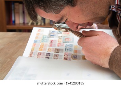 A Stamp Collector Checking His Philatelic Collection Of Stamps From Spain With A Magnifying Glass On A Wooden Table And A Library In A Blurred Background.  Jaen - Spain On 22nd Of March Of 2021