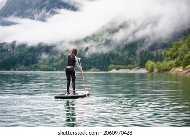 Stan up paddle female in wetsuit paddling on a big lake with green mountains and low white clouds on a foggy rainy day in Norway