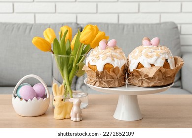 Stand with Easter cakes, painted eggs, bunnies and tulip flowers on table in living room