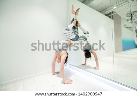 Stand near window. Dark-haired woman wearing leggings and top doing arm stand near window