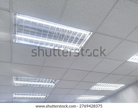 A standard suspended grid ceiling featuring recessed fluorescent light fixtures, illuminating an empty office space. The image provides a low-angle view of the textured ceiling tiles and the bright linear light tubes