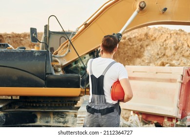 Standing against yellow vehicle. Worker in professional uniform is on the borrow pit at daytime.