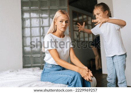 Standing angry little child daughter scolding, raising voice, yelling to sad depressed thinking young mother sitting on sofa at home. Concept of family problems, conflict, crisis.