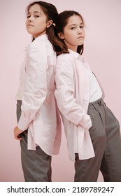 Standing Back To Back, Leaning On Each Other Twin Girls With Dress Shirt Over T Shirt. Wearing Pigtails And Black And White Patterned Pants Over Pink Background.