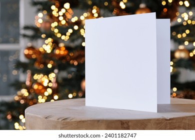 Standing blank empty square greeting card mock up before a blurred christmas tree with copy space. For use as a Christmas, background template.     