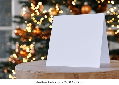 Standing blank empty square greeting card mock up before a blurred christmas tree with copy space. For use as a Christmas, background template.     