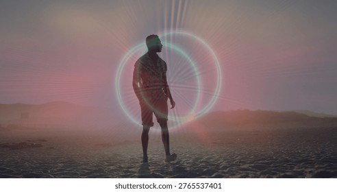 Standing lone man wearing patterned shirt and shorts on sandy beach, halo glowing, footprints. Silhouette, backlit, shoreline, horizon, twilight, pastel, coastal