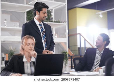 Standing manager checks progress over the woman's shoulder during an informal review, papers and coffee cups dotted between laptops in the coworking space in a bright modern coworking.