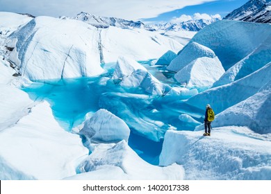 Standing Near The Edge Of Large Blue Pool On Top Of The Matanuska Glacier. A Young Woman Holding An Ice Axe With A Backpack And Helmet Looks Out Over The Lake.