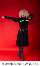Standing on tip toes Georgian ballet dancer man in classic costume. Bearded noble Georgian dancer in traditional Georgian costume and fur hat, posing on red-hot matte background