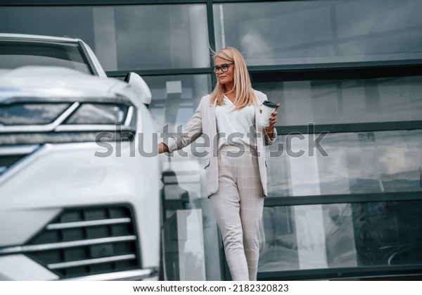 Standing outdoors\
against building and holding drink. Woman in white formal clothes\
is in the car\
dealership.