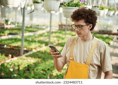 Standing, using smartphone. Young man with curly hair and in glasses is in greenhouse. - Powered by Shutterstock