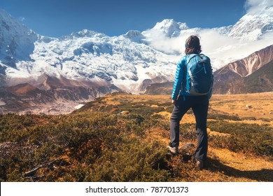 Standing young woman with backpack on the stone and looking on snow covered mountains in clouds at sunset. Landscape with girl, high rocks with snowy peaks, yellow grass, blue sky in Nepal. Travel