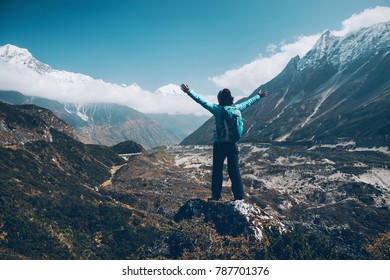 Standing young woman with backpack and raised up arms on the hill and looking on mountains. Landscape with happy girl, mountains, blue sky with clouds in autumn in Nepal. Travel. Trekking in Himalayas