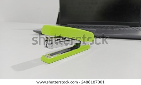 stapler on a white table and there is a laptop behind it. A stapler is a tool for holding a number of pieces of paper together.