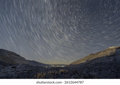 Star trails form a circular pattern over a snowy mountain valley at night, with snow-covered trees and distant lights illuminating the serene winter landscape.