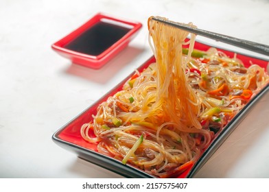 Starch Noodles With Vegetables And Beef In A Red Rectangular Plate On A Gray Background, Close-up. The Chopsticks Are Holding Starchy Noodles, There Is A Gravy Boat With Soy Sauce Next To It.