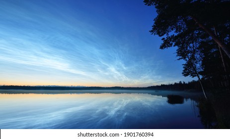 Starry Sky With Noctilucent Clouds And Fog Above The Saimaa Lake At Summer Solstice Night. Golden Sunlight. Long Exposure. Epic Cloudscape. Natural Pattern. Symmetry Reflections On The Water. Finland