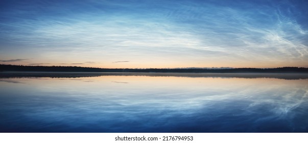 Starry sky with noctilucent clouds and fog above the Saimaa lake at summer solstice night. Tree silhouettes. Golden sunlight. Long exposure. Epic cloudscape. Symmetry reflections on the water. Finland - Powered by Shutterstock