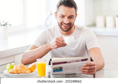 Starting day from good news. Handsome young man reading newspaper and smiling while drinking coffee and having breakfast in the kitchen - Powered by Shutterstock