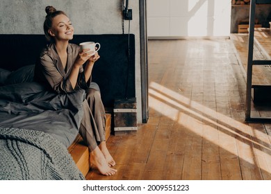 Starting New Beautiful Day. Cute Pretty Woman With Cup Of Morning Coffee Chilling While Sitting On Bed, Relaxing In Brown Satin Pajama In Modern Apartment. Happy Time At Home