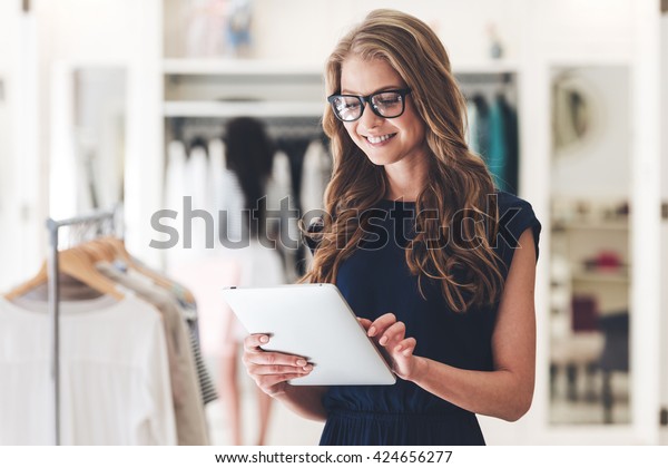 Starting new\
business. Beautiful young woman using digital tablet with smile\
while standing at the clothing store\
