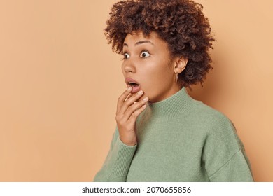 Startled curly haired woman holds chin and stares bugged eyes away holds breath from amazement notices something terrible wears casual jumper isolated over beige background blank copy space.
