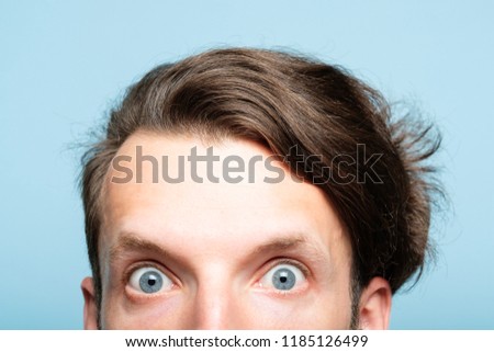 startled eccentric whimsical man with crazy look closeup. head of a young brunet guy peeking out from the bottom of blue background. emotion and facial expression concept.