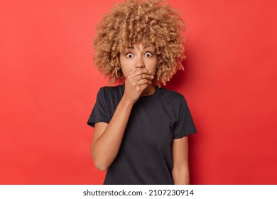 Startled frightened curly woman covers mouth with hand stares at something horrible tries to be speechless wears casual black t shirt isolated over vivid red background. Omg dont drop any word