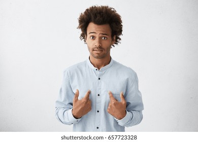 Startled handsome African businessman in elegant white shirt pointing at himself with hands trying to defence himself raising his eyebrows with great surprise. Man asking something about himself