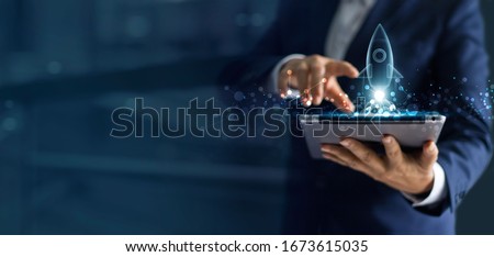 Startup business concept, Businessman holding tablet and icon transparent rocket is launching and soar flying out from screen with network connection on dark blue modern background.