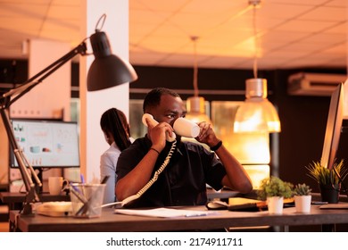 Startup employee using landline phone to have remote conversation during sunset. Chatting on office telephone with colleagues to work on business report and do paperwork contract. - Shutterstock ID 2174911711