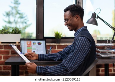 Startup Owner Holding Clipboard With Annual Bussiness Charts Happy With Turnover Results Sitting At Desk With Laptop. Entrepreneur Comparing Key Performance Indicators On Portable Computer.