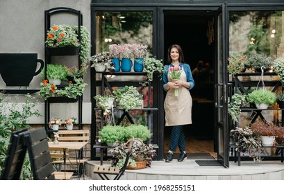 Startup, Small Business, Eco Restaurant Outdoor And Modern Rustic Flower Shop. Smiling Millennial Beautiful Female In Apron Holds Bouquet Of Tulips For Client, At Front Door Of Plants Studio Or Cafe