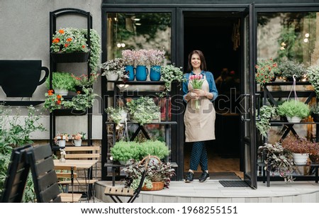 Startup, small business, eco restaurant outdoor and modern rustic flower shop. Smiling millennial beautiful female in apron holds bouquet of tulips for client, at front door of plants studio or cafe