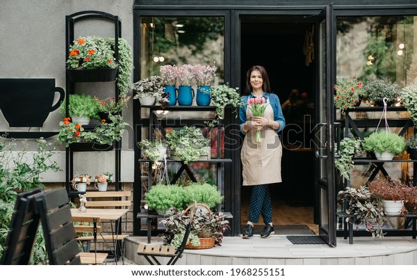 Startup, small business, eco restaurant outdoor and
modern rustic flower shop. Smiling millennial beautiful female in
apron holds bouquet of tulips for client, at front door of plants
studio or cafe